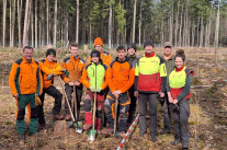 Eine Gruppe von Menschen in Orangefarbenen Jacken stehen auf einer kahlen Fläche im Wald und halten Spaten in den Händen