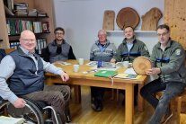 5 Männer, davon einer im Rollstuhl sitzen rund um einen Tisch, ein Mann hält eine Baumscheibe in der Hand
